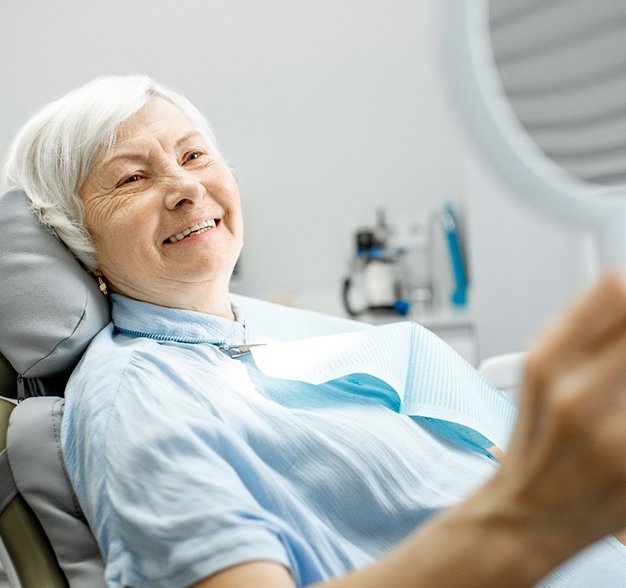 An older woman seated in the dentist’s chair admiring her new smile