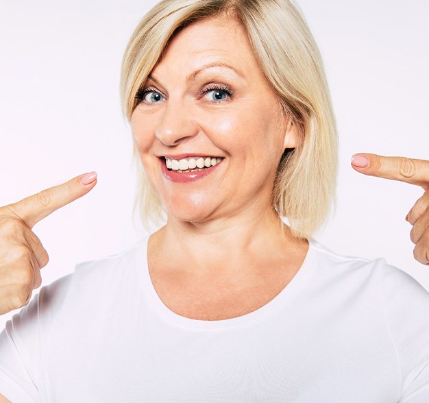 An older woman wearing a white t-shirt and pointing to her new and improved smile, complete with dental implants